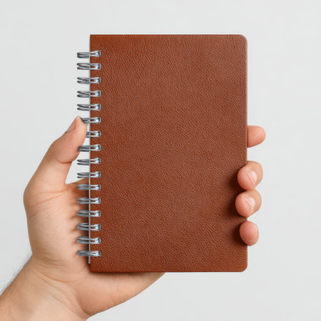 A hand presents a small brown spiral notebook featuring a textured cover against a plain backdrop, ideal for writing and organizing thoughts.の素材
