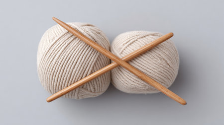 Two soft cream colored yarn balls and wooden knitting needles arranged in a creative flat lay on a gray background, showcasing cozy crafting supplies.の素材