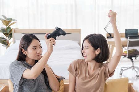 Two Asian girlfriends enjoy playing video games with joysticks at home. Lesbian couples or lgbt...