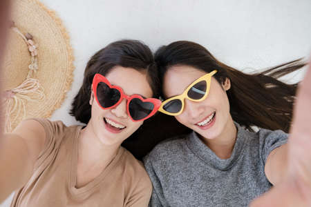 Portrait of two young Asian women wearing sunglasses in summer concept smiling and lying on the white floor to selfy take a photo.の写真素材
