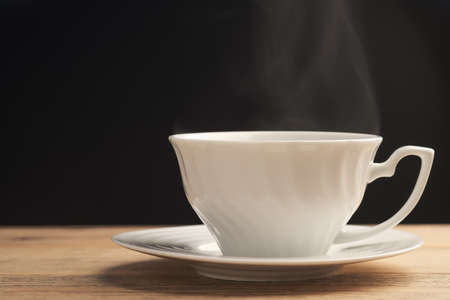 A white cup of coffee or tea with smoke on old wooden table on black background. Hot drink.