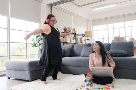 Asian mother using laptop and young girl with autism wearing headphone dancing on the floor at home with his mother. Autistic young students are learning with teachers.の写真素材