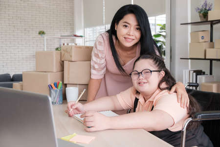 Middle-aged mother with Asian autistic daughter is looking at the camera and smiling inside home converted into small business office online. New business in the era of the COVID-19 epidemic.の写真素材