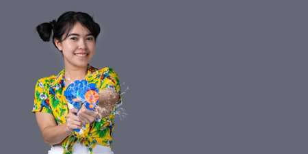 Portrait cheerful young Asian woman holding water gun Smiling and having fun playing in the water Songkran festival, Thailand. isolated on grey background. Thai New Year's Day.の写真素材