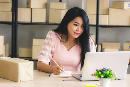 Asian businesswoman start-up online seller owner using computer for checking the customer orders from email or website and preparing packages - online shopping or sell online conceptの写真素材