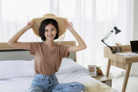 Young Asian woman smiling and experimenting with hats and packing suitcases for summer vacation travel in his bedroom. concept of travel in summer and holiday.の写真素材