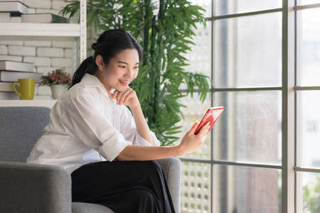 young Asian woman relaxing on sofa inside the house. Young women use tablet to entertainment Communicate and transact online. work from home.の写真素材