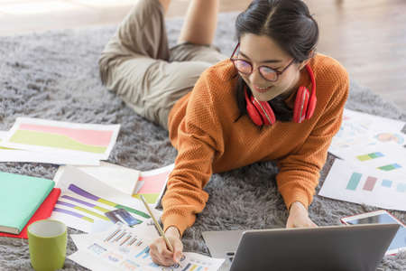 A young Asian business woman sits at home using a laptop computer operated by a video conferencing...