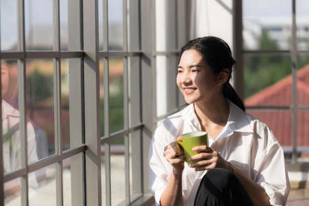 Young Asian woman smiling and drinking coffee by the window with the morning sunshine at home.