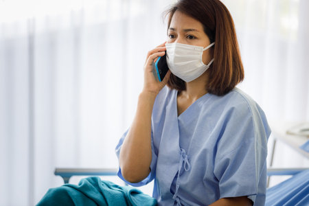 A middle-aged Asian female patient wearing a mask is talking on the phone with a smartphone in bed in a hospital. quarantine and social distancing of the outbreak of the COVID 19 virusの写真素材