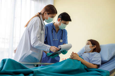 Asian female and male doctors wearing masks and uniforms are visiting to check the symptoms of middle-aged female patients lying in bed in the hospital. quarantine and social distancing of the outbreak of the COVID 19 virus.の写真素材