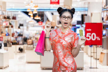 Portrait of beautiful Asian woman wearing red cheongsam and carrying shopping bag with credit card....