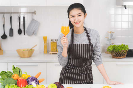 Portrait of beautiful asian woman, housewife with orange juice in white kitchen with plenty of fresh vegetables and fruits on the table.の写真素材