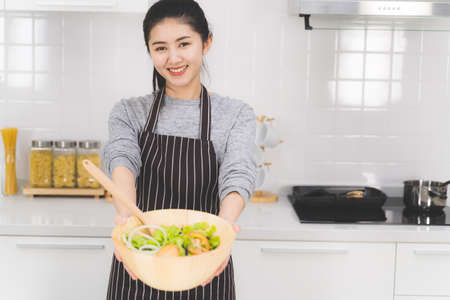 Portrait of beautiful Asian woman wearing an apron smiling and holding a bowl of salad. Healthy food concept.の写真素材