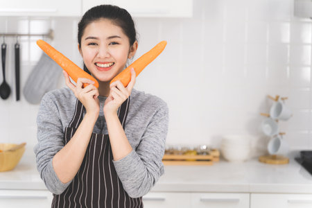 Portrait of asian beautiful woman and housewife is smiling and holding carrots in white kitchen. Healthy food concept.の写真素材