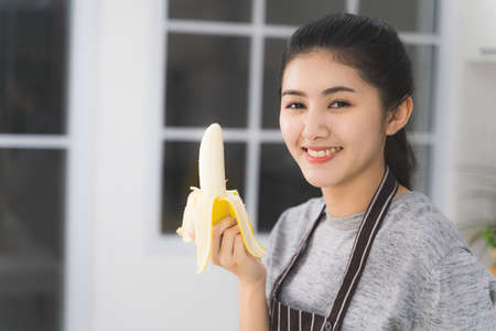 Portrait of asian beautiful woman and housewife is smiling and holding banana in white kitchen. Healthy food concept.の写真素材