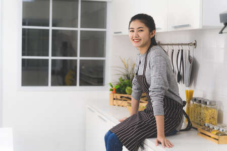Portrait of asian woman, housewife smiling and sitting on counter in white kitchen.の写真素材