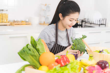 Beautiful asian woman is preparing to cook healthy food which consists of a variety of fruits and vegetables for the family. Housewife is smiling and holding broccoli.の写真素材