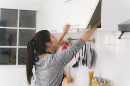 Beautiful asian woman, housewife is searching for something from the cabinet above the kitchen counter.の写真素材