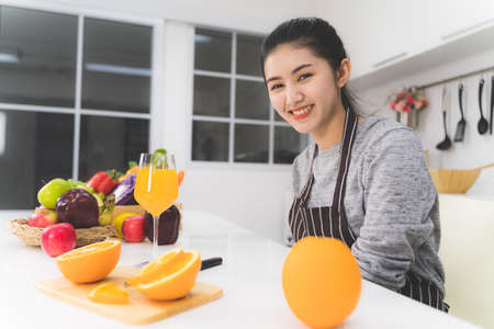 Portrait of beautiful asian woman, housewife with orange juice in white kitchen with plenty of fresh vegetables and fruits on the table.の写真素材