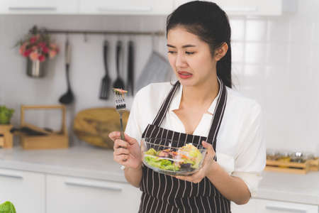 Beautiful Asian woman is feeling terrible with a taste of food and shows a fork that cucumbers from a salad bowl in a white kitchen with lots of fresh vegetables and fruits on the table.の写真素材