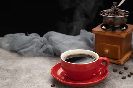 Red cup of black coffee with smoke, grinder and coffee beans on cement table.の写真素材