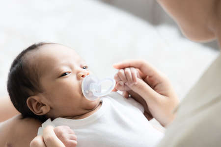 Portrait of cute newborn baby sucking fake nipples and looking hers mother in white bedroom. Duty of the wife to raise children. Mother and child conceptの写真素材