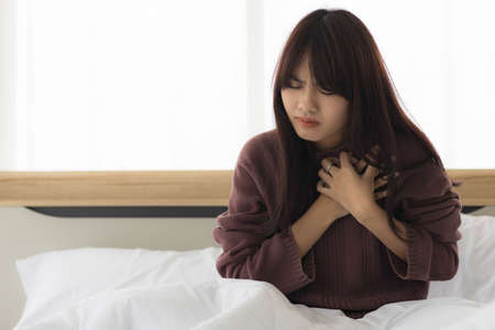 Asian woman in her bedroom uses her hand to hold her heart due to the aggravated heart disease....