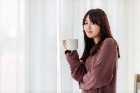 Asian woman smiling and holding cup of coffee in her bedroom. corovavirus covid-19, stay at home, work form home concept.の写真素材