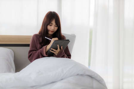 Asian woman using tablet for working, educational, e-commerce, social media in her bed. corovavirus covid-19, stay at home, work form home concept.の写真素材
