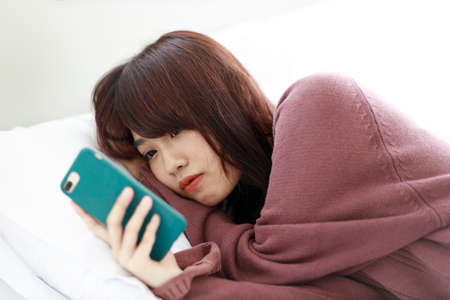 Asian woman watching media from phone in a bedroom in her bedroom feeling lonely. The concept of daily activities of people in the capital and housewives.の写真素材