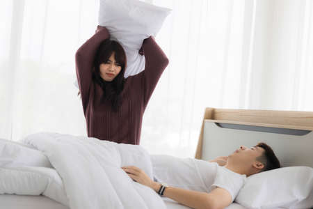 Asian wife is using a pillow to hit her husband because she is irritated by her snoring. The concept of married life problems after marriageの写真素材