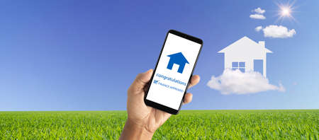 Smartphone mobile phone that approves home loan on paper house on clouds against meadow background sky and the sun background. Dream home. Loan, mortgage, and real estate concepts.の写真素材