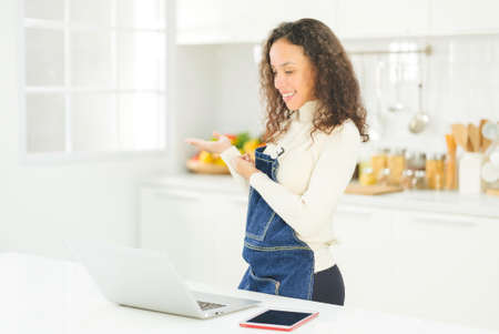 Portrait of beautiful young black woman is live streaming cooking with laptop in the bright white kitchen. wife or housewife is preparing to cook healthy food for the family. Stay at home.の写真素材