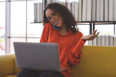 Beautiful young black woman wearing orange shirt using laptop and smartphone for communication on sofa at home. Business woman and freelance work from home in spreading of coronavirus covid-19.の写真素材