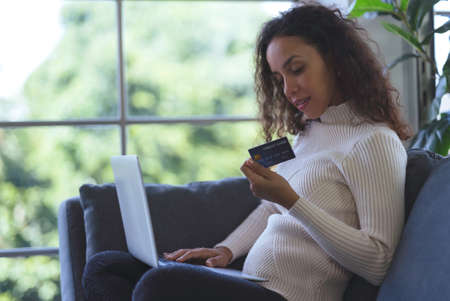 Beautiful young black woman smiling and using laptop and credit card for shopping online on sofa at home. Business woman and freelance work from home in spreading of coronavirus covid-19.の写真素材