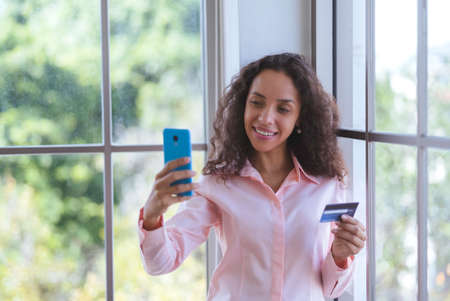 Young latin woman smiling and using smartphone for online shopping. Business woman and freelance work from home and holding credit card for electronic payment. happy shopper services in online store.の写真素材
