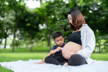 Portrait of a pregnant woman relaxing in the park with her son. The older brother uses a stethoscope to listen to the sound of his younger brother in his mother's womb.の写真素材