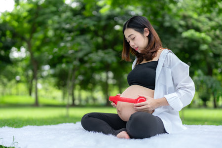 Portrait of Asian pregnant woman relaxing and in the park. Young woman uses headphones on her stomach to let her fetus listen to music.の写真素材