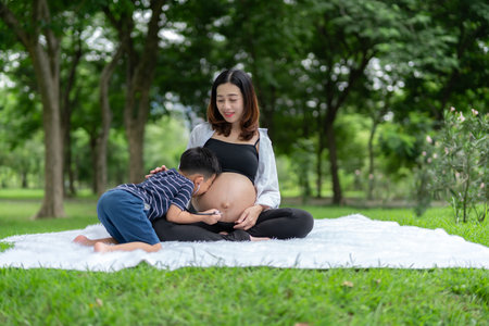 Portrait of a pregnant woman relaxing in the park with her son. The older brother uses a stethoscope to listen to the sound and kiss of his younger brother in his mother's womb.の写真素材