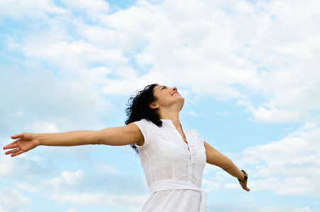 Happy smiling woman standing with outspread arms and her face lifted to the cloudy blue skyの写真素材