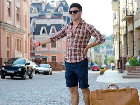 Young male hitchhiker standing with his suitcase in an urban street thumbing a liftの写真素材