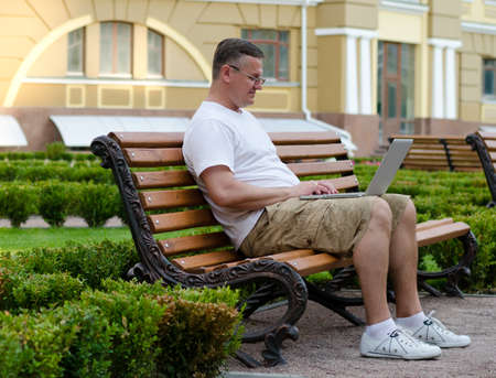 Man sitting in front of an urban building using a laptop on a public bench in a manicured gardenの写真素材
