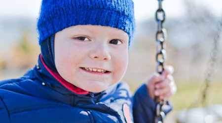 nice little boy on a swing in a jacket and a cap in the autumn, smiles and looks in the chamber