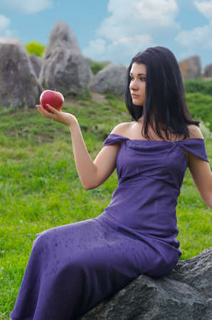 Elegant beautiful woman posing with a red apple balanced on the palm of her hand sitting on a rock outdoorsの写真素材
