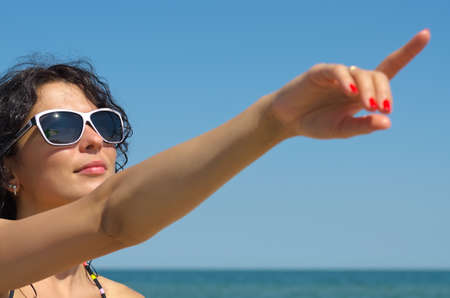 Beautiful woman in sunglasses pointing with an extended arm to the blue sky above her headの写真素材