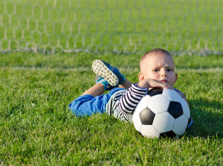 Cute young boy with a frown on his face lying on grass with a soccer ball as he waits for someone to play with himの写真素材