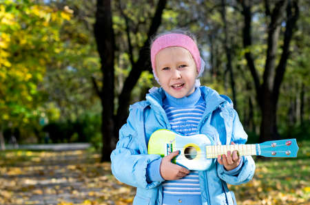 Happy little girl standing outdoors alone in autumn woodland strumming a blue toy guitarの写真素材