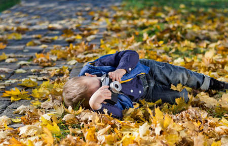 Small boy photographng Autumn lying in a pile of colourful yellow fall leaves with his vintage slr film cameraの写真素材