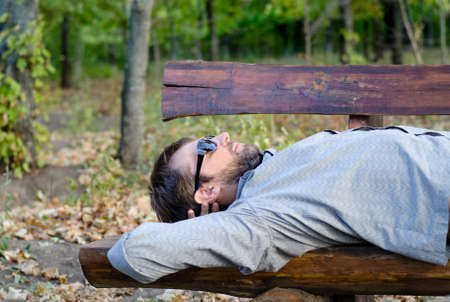 Man sleeping on a rustic wooden bench lying back on the seat in the sunshine with his sunglasses onの写真素材